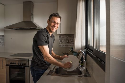 Smiling and happy man washing dishes in the kitchen next to a window. Medium shot.