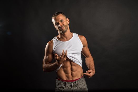Athletic man lifting his shirt to show his abs. Mid shot. Black background.