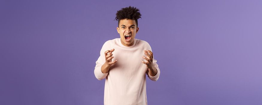 Portrait of hateful, outraged young hispanic man losing his temper, want to strangle or attack someone, cursing being aggressive, squeeze hands into fist from rage and hate, purple background.