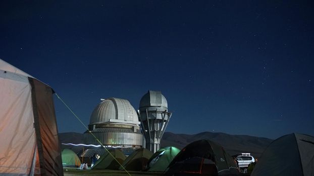 A camp with colored tents near the observatory. The Perseid starfall. A large teleskom watches the starry night sky. Meteorites and planes fly by, leaving traces in the sky. The stars are burning.