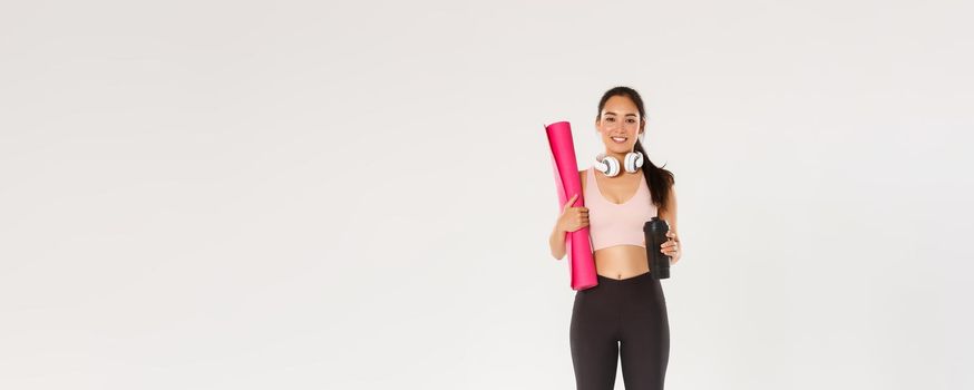 Full length of cute brunette asian fitness girl with headphones, activewear and water bottle, carry pink rubber mat for gym exercises, going for yoga classes, standing over white background.