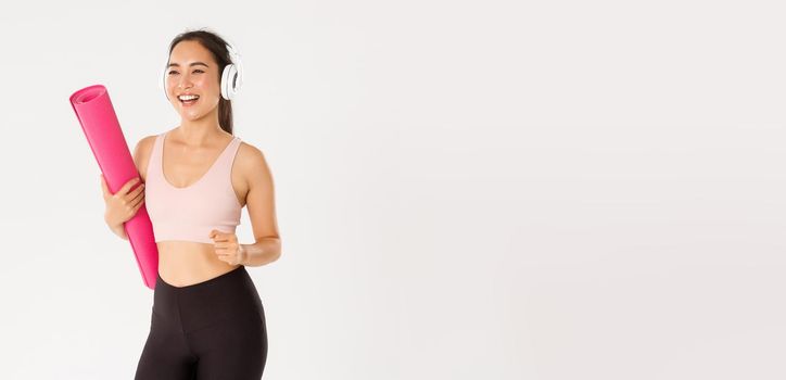 Sport, wellbeing and active lifestyle concept. Cheerful smiling asian fitness girl in headphones and sportswear going to gym with rubbermat, hurry up for yoga classes excited, white background.