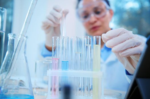 Selective focus on test tubes with chemical fluid substances, next to laboratory glassware over blurred scientist pharmacologist or lab technician, making researches and analyzes in medicine field.
