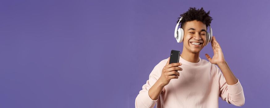 Technology and lifestyle concept. Portrait of carefree attractive young man enjoying fine sound, listening music with headphones look delighted awesome quality of bass, hold mobile phone.