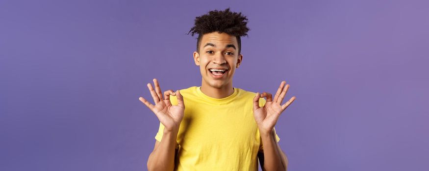 Close-up portrait of upbeat, relaxed young hipster guy with dreads assuring all good, everything be okay, show OK gesture and smiling, no problem guarantee best quality, purple background.