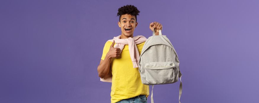 Portrait of cheerful young handsome male student, man recommending backpack, holding rucksack and show thumbs-up, bought new equipment for university semester, purple background.