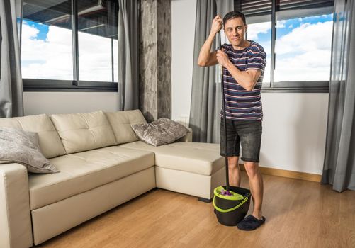 Smiling man in the living room wearing casual clothes and slippers, wringing the mop into the bucket. Full body.