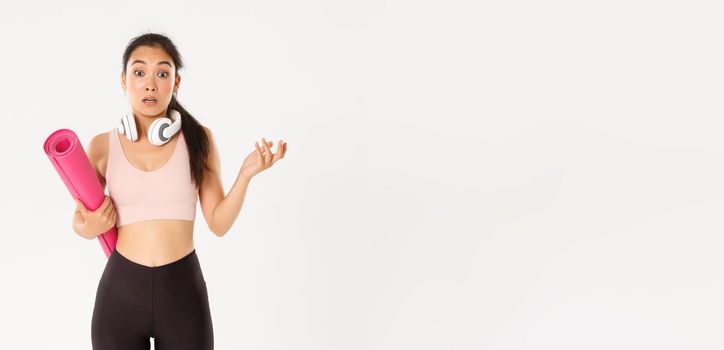 Sport, wellbeing and active lifestyle concept. Confused and shocked asian fitness girl, cant understand why gym closed, holding rubber mat and looking surprised at camera, white background.