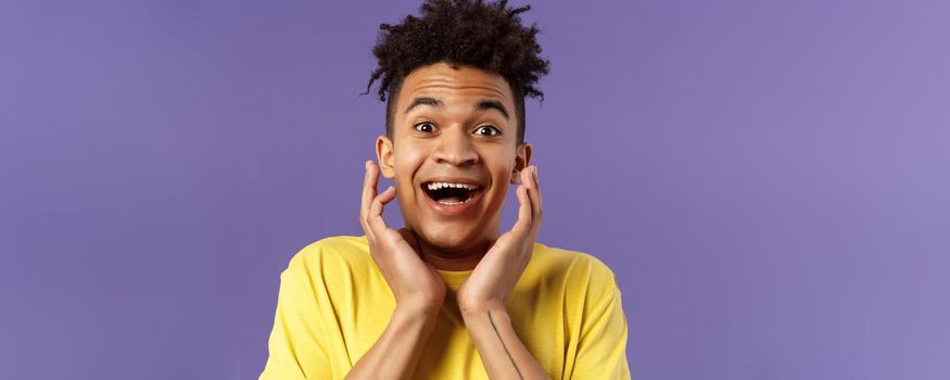 Close-up portrait of extremely happy, enthusiastic young man hear fantastic news, looking surprised and excited, touching face in joy, smiling upbeat look camera astonished, purple background.