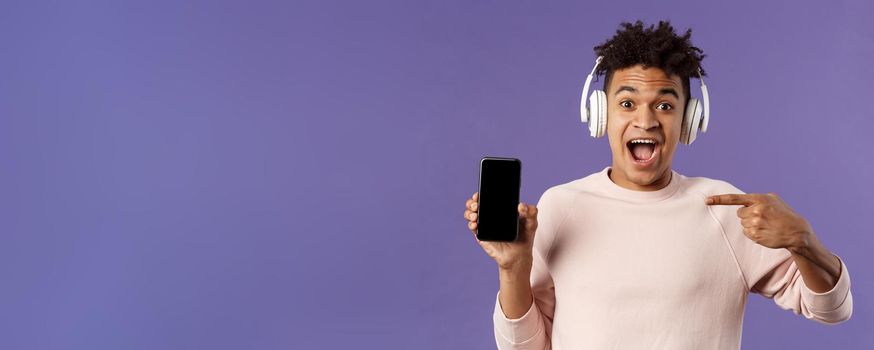 Technology and lifestyle concept. Portrait of excited impressed young man in headphones listening music, online platform or enjoying interesting podcast, pointing at mobile phone, recommending.