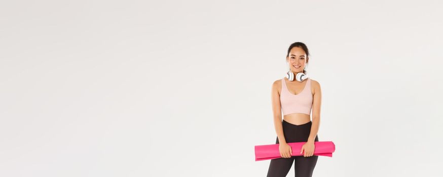 Full length of smiling slim asian girl with rubber mat for fitness exercises, headphones, looking at camera while waiting for training coach starting workout session in gym, white background.