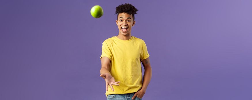 Holidays, vitamins and vacation concept. Portrait of handsome upbeat young male student asking friend something eat, catching apple and smiling happy, standing purple background.
