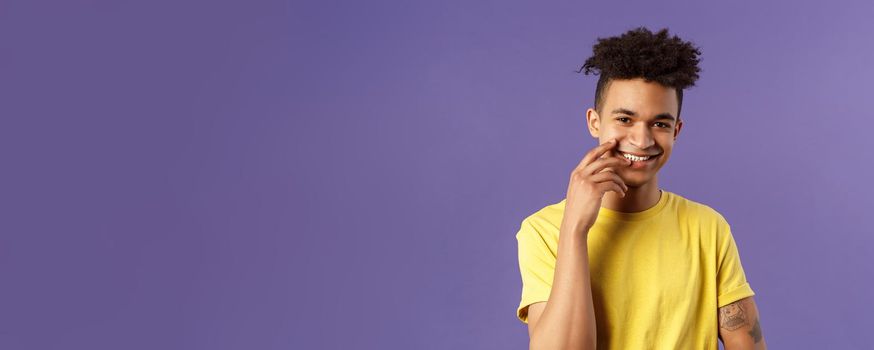 Close-up portrait of lovely young hispanic gay man with dreads, tattoos, touching lip sensually and flirty smiling, checking out someone really cute and handsome, standing purple background.