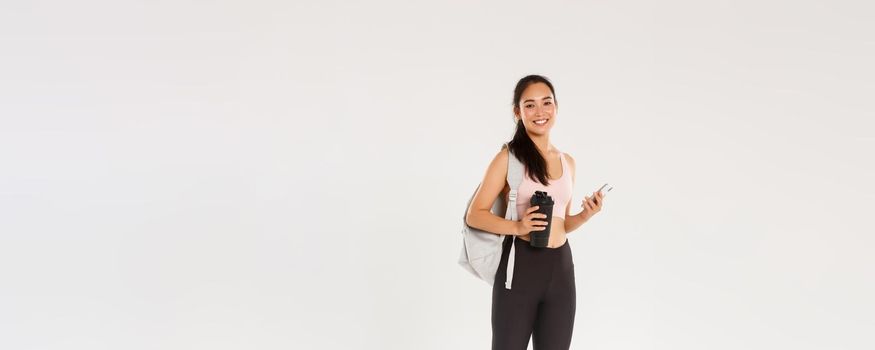 Full length of smiling healthy, slim asian girl going fitness training, female athelte carry backpack with workout equipment and water bottle, using mobile phone sports application, white background.