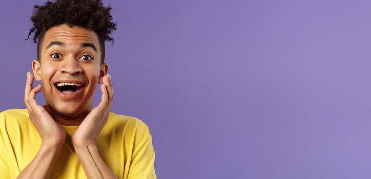 Close-up portrait of extremely happy, enthusiastic young man hear fantastic news, looking surprised and excited, touching face in joy, smiling upbeat look camera astonished, purple background.