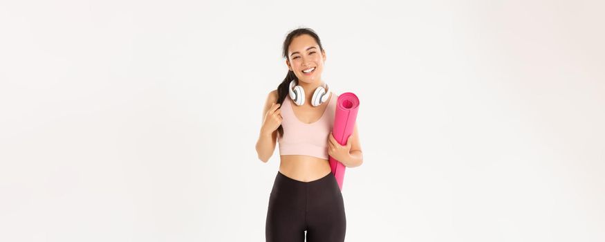 Sport, wellbeing and active lifestyle concept. Smiling happy asian fitness girl in headphones and sportswear, carry rubber mat for workout or yoga, laughing carefree over white background.