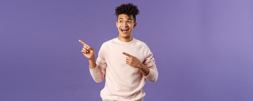 Portrait of upbeat, good-looking hispanic man with dreads, smiling amused, looking and pointing upper left corner, advertisement of product or company banner, copy space, purple background.
