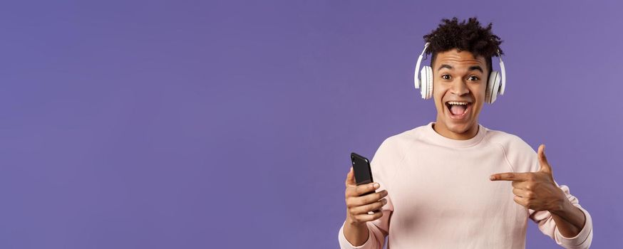 Technology and lifestyle concept. Portrait of happy, cheerful young man recommend awesome podcast or online music platform, buying subscribtion listen songs anytime, wear headphones, point at phone.