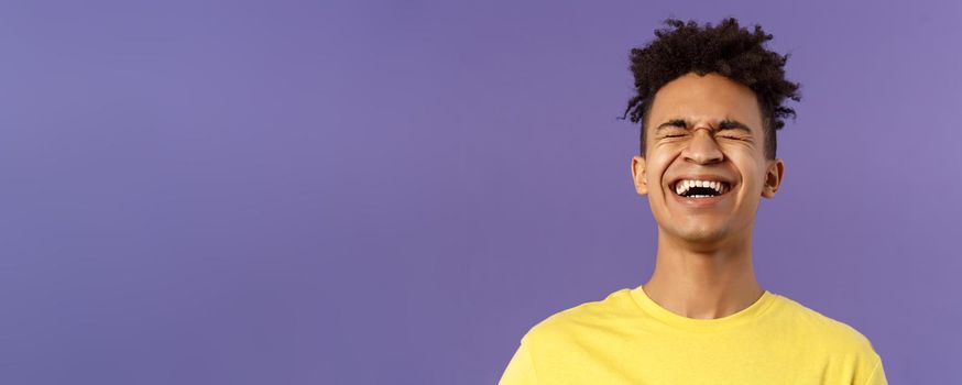 Close-up portrait of happy carefree young guy laughing loud, chuckling over hilarious joke, bending backwards and close eyes while giggle over funny movie, purple background.