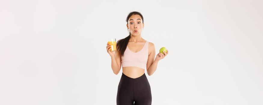Sport, wellbeing and active lifestyle concept. Amused and wondered cute asian girl likes fitness and eating healthy food, holding apple and orange juice, looking amazed saying wow, white background.