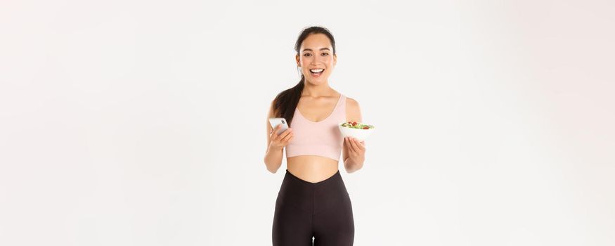Sport, wellbeing and active lifestyle concept. Smiling slim asian fitness girl in sportswear, holding salad and mobile phone, using eating reminder app, diet control application, checking calories.