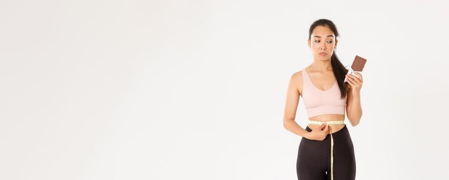 Sport, wellbeing and active lifestyle concept. Thoughtful and indecisive asian girl looking gloomy over body weight, measuring waist but looking at chocolate bar, pondering, white background.
