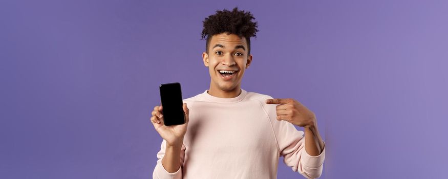 Portrait of excited, happy smiling man talking about new application or game feature, pointing at smartphone display, talking about mobile phone with amused cheerful expression.