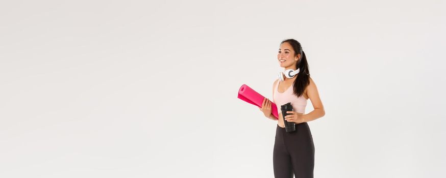 Full length side view of beautiful asian girl going for yoga classes, female athlete with headphones and water bottle smiling as carry fitness exercises rubber mat for her training in gym.