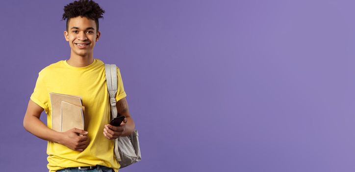 Back to school, university concept. Portrait of young cheerful male student with dreads, hipster going to his campus, carry backpack and study material, notebooks, purple background.
