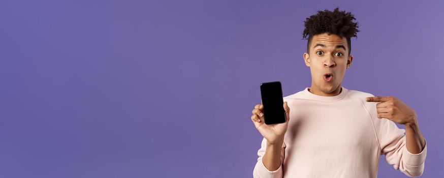 Close-up portrait of excited, cheerful young hispanic male geek talking about his device, new gadget or application, holding mobile phone, pointing at smartphone with amused face.