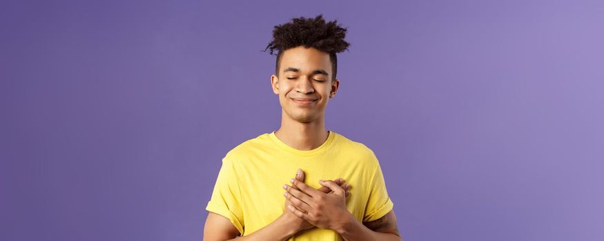 Always in my heart. Touched, carefree handsome young man with dreads, yellow t-shirt, close eyes and smiling, hold hand on chest as recall memory of his first love, feel heartwarming emotion.