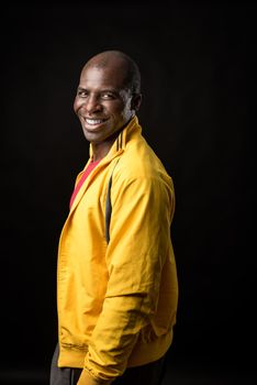 Side view of an African American man standing looking at camera and smiling on black background. Cheerful adult male in yellow jacket and red t-shirt in a studio with black background.