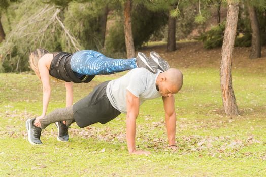 Fitness couple doing push ups on the back in the park. Multi-ethnic people exercising outdoors.