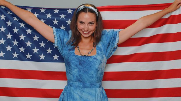 Lovely young woman in blue dress waving and wrapping in American USA flag, celebrating, human rights and freedoms. Independence day. Adult stylish girl isolated alone on gray studio background indoor