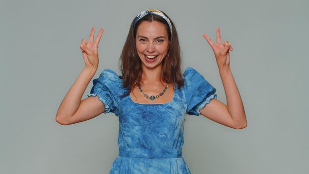 Lovely happy young woman showing victory sign, hoping for success and win, doing peace gesture, smiling with kind optimistic expression. Adult stylish girl isolated on gray studio background indoors