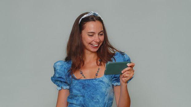 Worried funny young woman in blue dress enthusiastically playing racing or shooter video games on smartphone. Stylish girl using smartphone gadget app with drive simulator on gray studio background