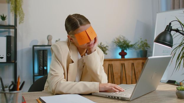 Inefficient tired businesswoman in suit working sleeping on laptop computer with eyes stickers on face at office workplace desk. Funny lazy manager freelancer woman. Business people. Cheating to sleep