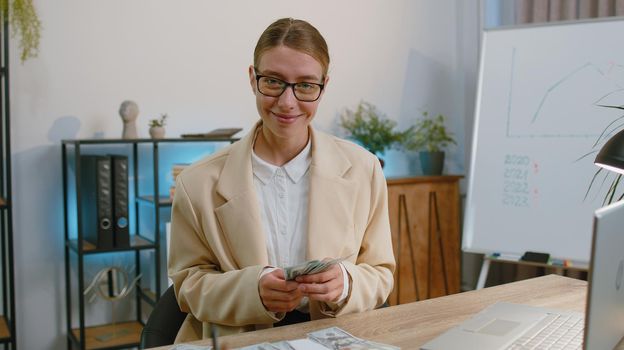 Rich happy businesswoman in suit counting money cash, calculate financial bills use laptop computer working at office workplace desk. Freelance woman satisfied of income earnings, profit. Girl worker