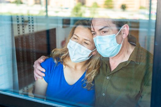 Mature couple with face mask looking sad out the window from home. High quality photo