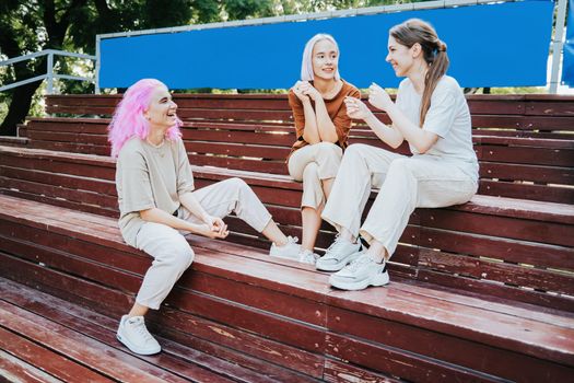 Modern teenage girls with colorful dyed hair sitting on bench in park. Women chatting, gossiping and laughing. Friendship concept. High quality photo
