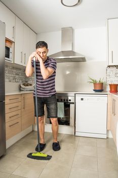 Sad and pensive man wearing casual clothes and slippers sweeping the kitchen floor. Full body.