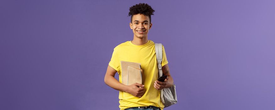 Back to school, university concept. Portrait of young cheerful male student with dreads, hipster going to his campus, carry backpack and study material, notebooks, purple background.