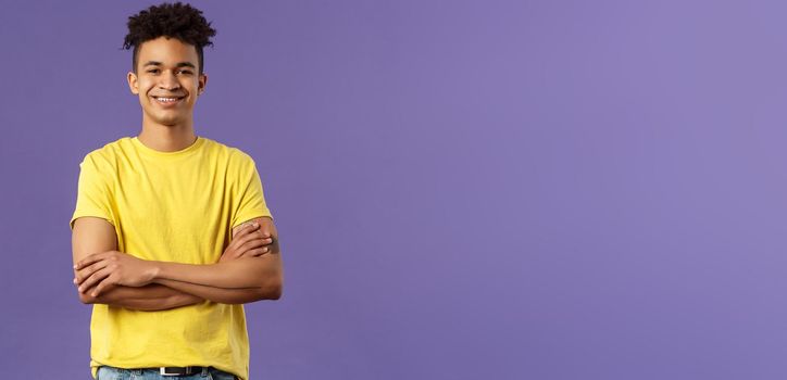 Close-up portrait of confident, smart and professional young male student with dreads, yellow t-shirt, cross arms over chest and smiling pleased, knows what he doing, purple background.