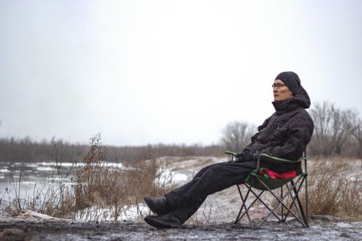 Resting brooding asian man sit on camping chair by frosty river in november, cold cloudy overcast weather in late autumn or winter with first snow