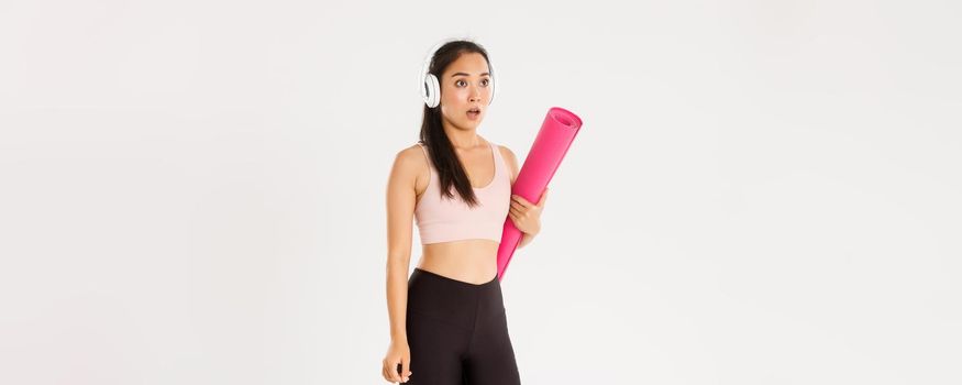 Sport, wellbeing and active lifestyle concept. Shocked and startled asian girl in headphones, holding rubber mat, stop and drop jaw as looking at logo or closed sign, covid-19 quarantine measures.