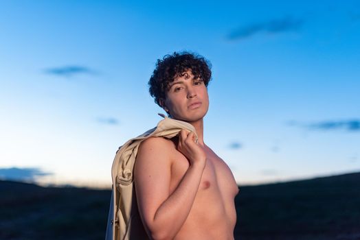 Portrait during dusk of a non binary male with naked torso facing the camera and holding a shirt