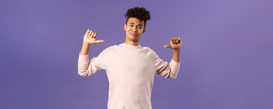 Portrait of boastful handsome young male student with dreads, bragging about how he workout in gym, show-off, pointing at himself and smirk self-assured, standing purple background.