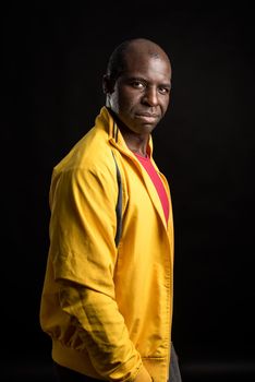 Side view of an African American man standing looking at camera. Adult male in yellow jacket and red t-shirt in a studio with black background.