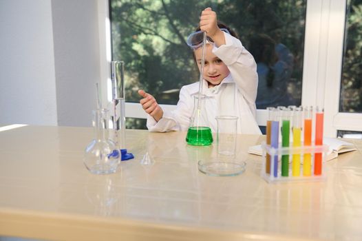 Little schoolgirl in safety goggles and white lab coat, student in chemistry class, conducts scientific experiments in the school laboratory. Back to school. Child scientist study science.
