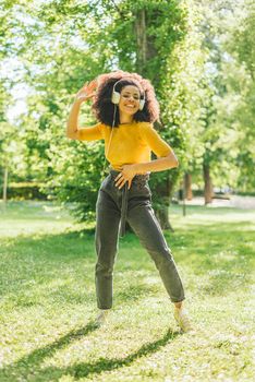 Pretty afro woman listens to music with headphones dancing in a garden. Selective focus.